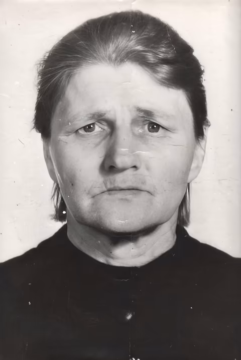 Черно-белая портретная фотография Анны Алексеевны Осиповой, ветерана Великой Отечественной войны. Пожилая женщина с достойной выдержкой смотрит прямо в объектив камеры с серьёзным, задумчивым выражением лица. Её тёмные волосы аккуратно разделены пробором по центру и убраны назад, открывая лицо, изборождённое глубокими морщинами и складками, которые свидетельствуют о десятилетиях стойкости и невзгод. На ней тёмная одежда с высоким воротником, характерная для советской формальной одежды. Винтажная фотография несёт следы времени — тонкие царапины, едва заметные заломы и выцветание, — что придаёт ей подлинный исторический характер и указывает на её происхождение из середины XX века.