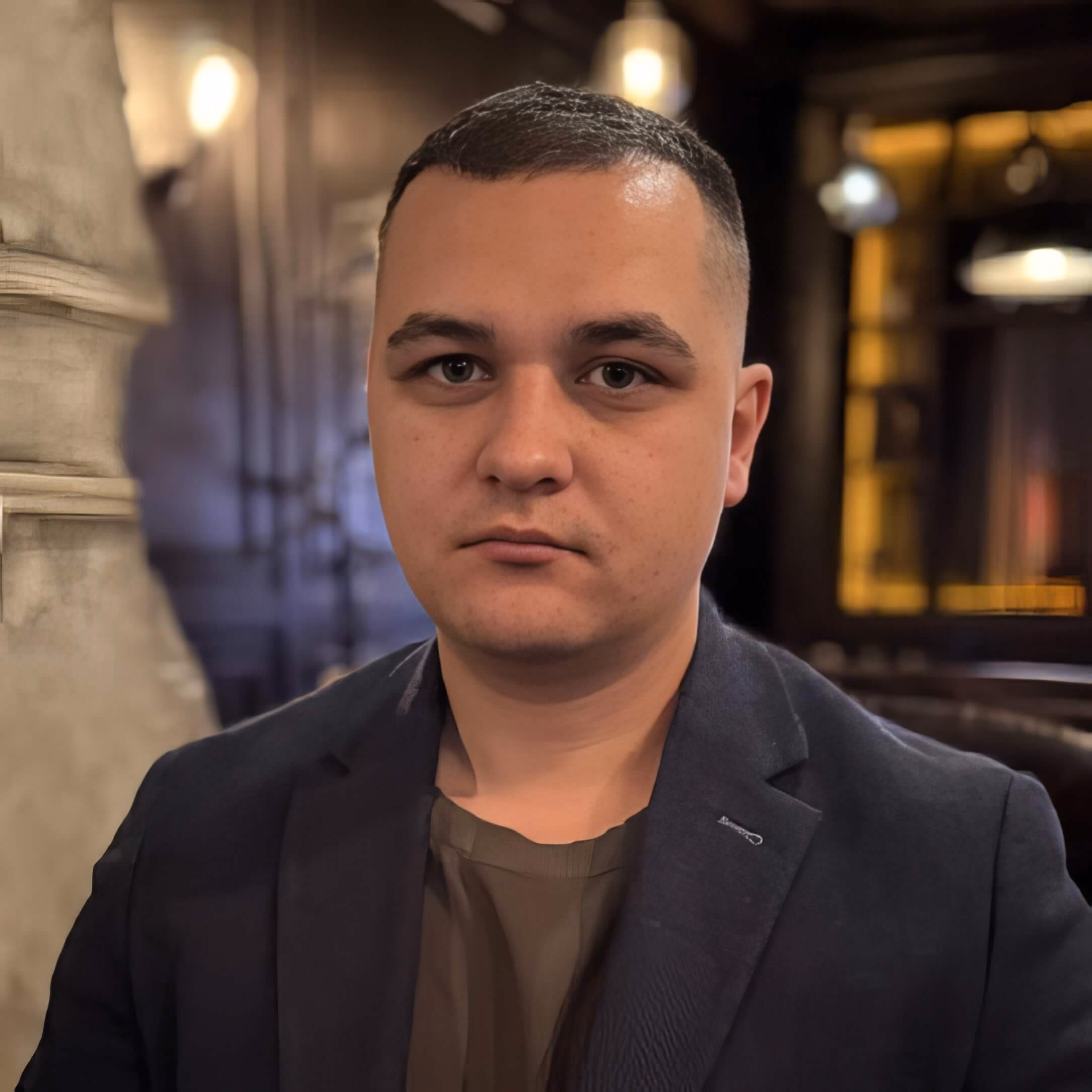 Крупный портрет Артема Гяммера, молодого мужчины с округлым лицом и светлой кожей. У него короткие темно-каштановые волосы: сверху они чуть длиннее, уложены вперед (стрижка кроп), а виски коротко выбриты с плавным переходом (фейд). Лоб широкий и гладкий. Брови густые, темные, естественной формы, слегка сведены к переносице, что придает взгляду серьёзность и сосредоточенность. Глаза темные, глубоко посаженные, миндалевидной формы, смотрят прямо в объектив. Нос прямой с округлым кончиком и слегка широкими крыльями. Губы сомкнуты, выражение лица нейтральное; верхняя губа тоньше нижней и имеет мягкий изгиб ('лук Купидона'), нижняя губа более полная. Линия челюсти мягкая, подбородок округлый. Лицо гладко выбрито, но над верхней губой и на подбородке заметна очень легкая тень от щетины. На правой щеке (слева для зрителя) видна небольшая родинка. Одет в тёмно-оливковую майку и темный блэйзер.